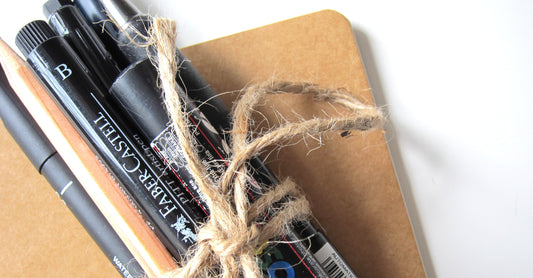 A bundle of paint markers and pens laying on a kraft-colored notebook.