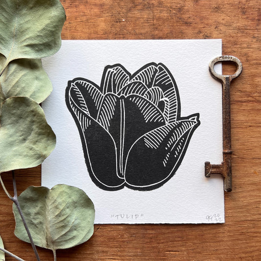 A 4x4” linocut print of a tulip printed in black ink on white paper. The linocut is laying on a wood surface with a eucalyptus branch to the left of the linocut.