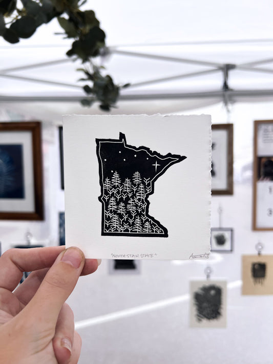 A hand holding a "North Star State" print up against a wall full of other linocut prints.