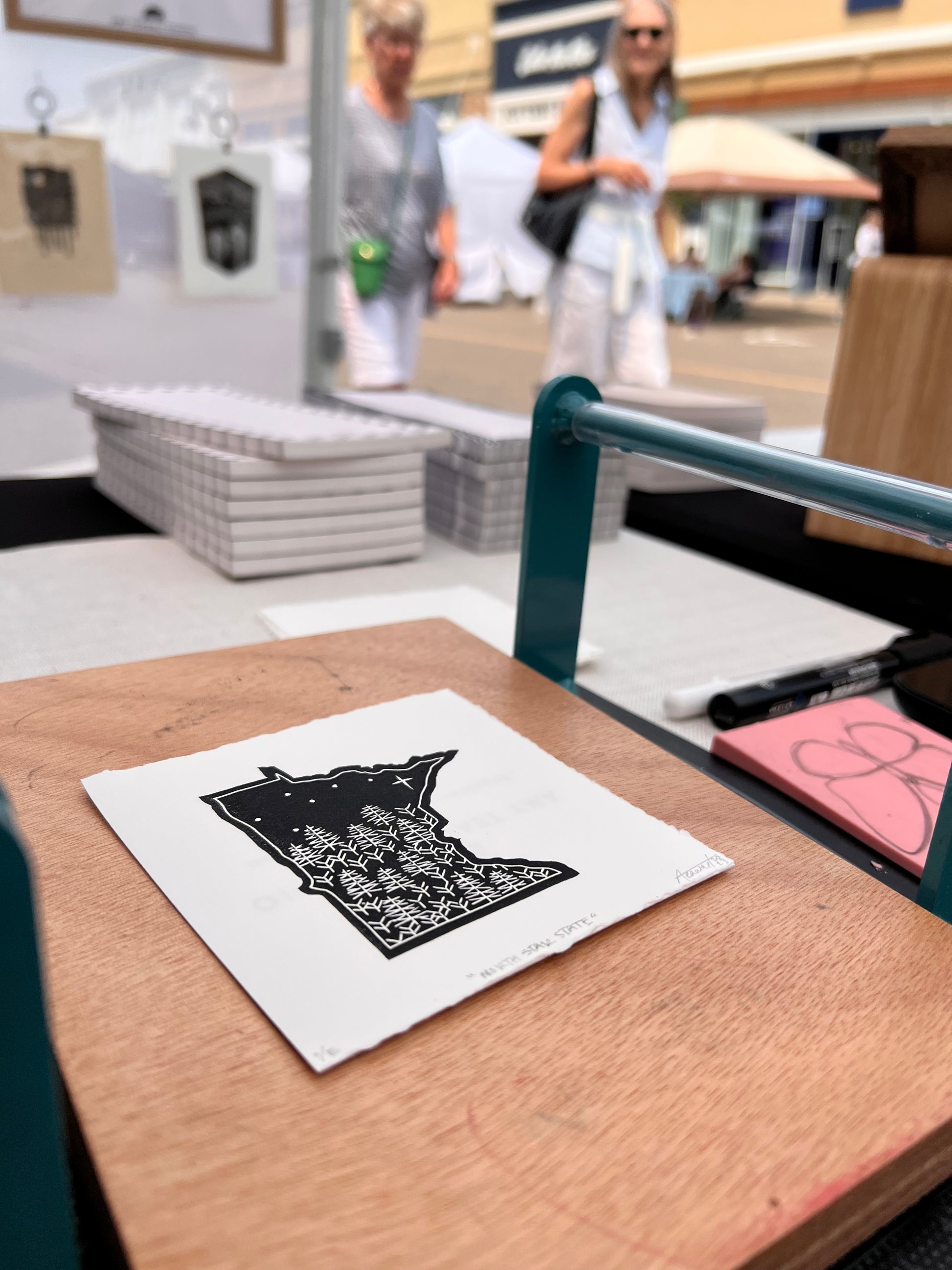 A "North Star State" mini print sitting on a tabletop press. In the background are other parts of the tablescape and people walking by on the other side of the table.